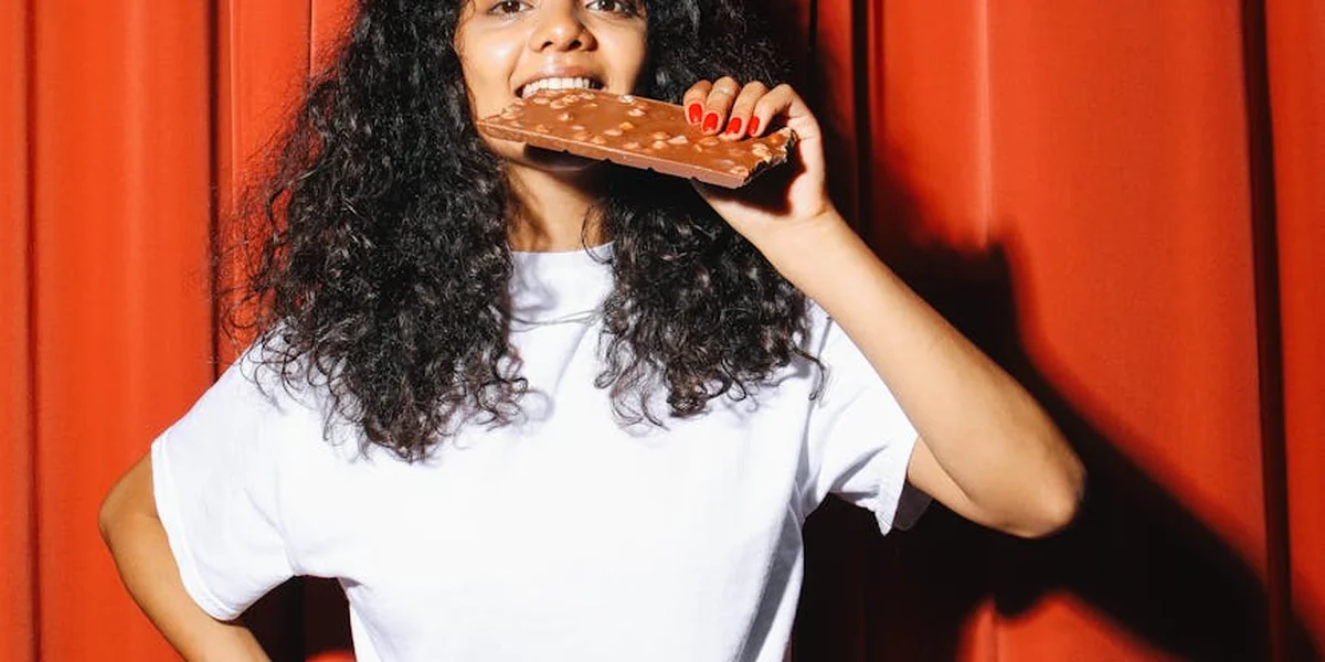 Person with curly hair biting into a chocolate bar against a bold red background.