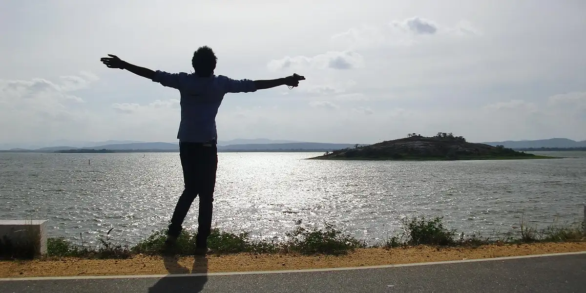 Silhouette of a person standing on a roadside by a body of water with arms outstretched against the sky.