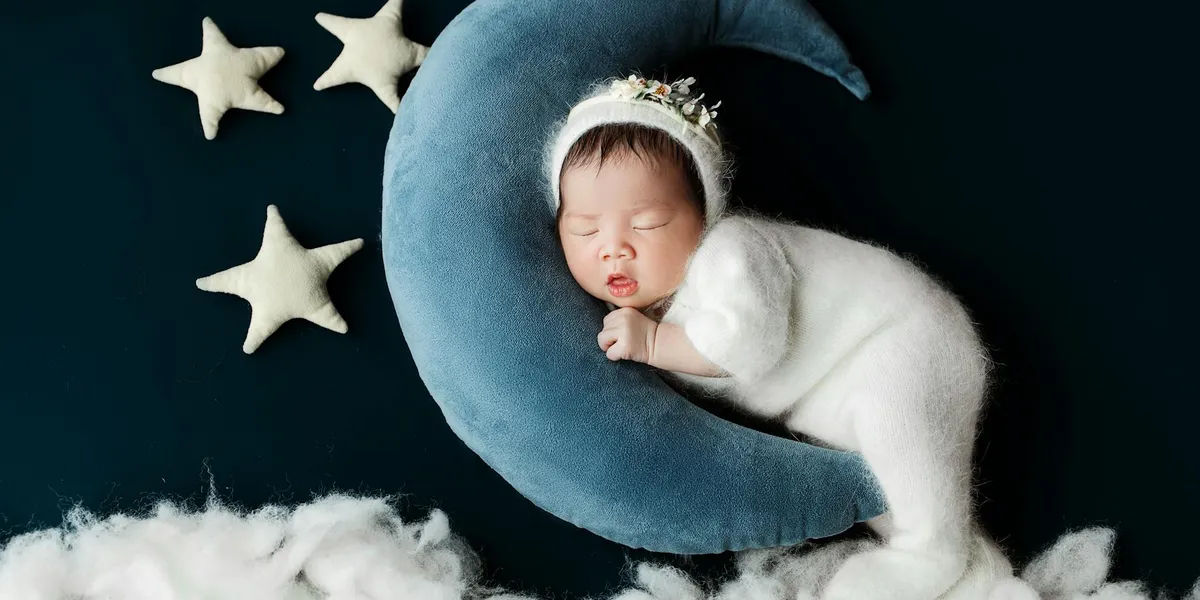 A sleeping baby wearing a crown rests on a blue crescent moon with white stars against a dark backdrop.