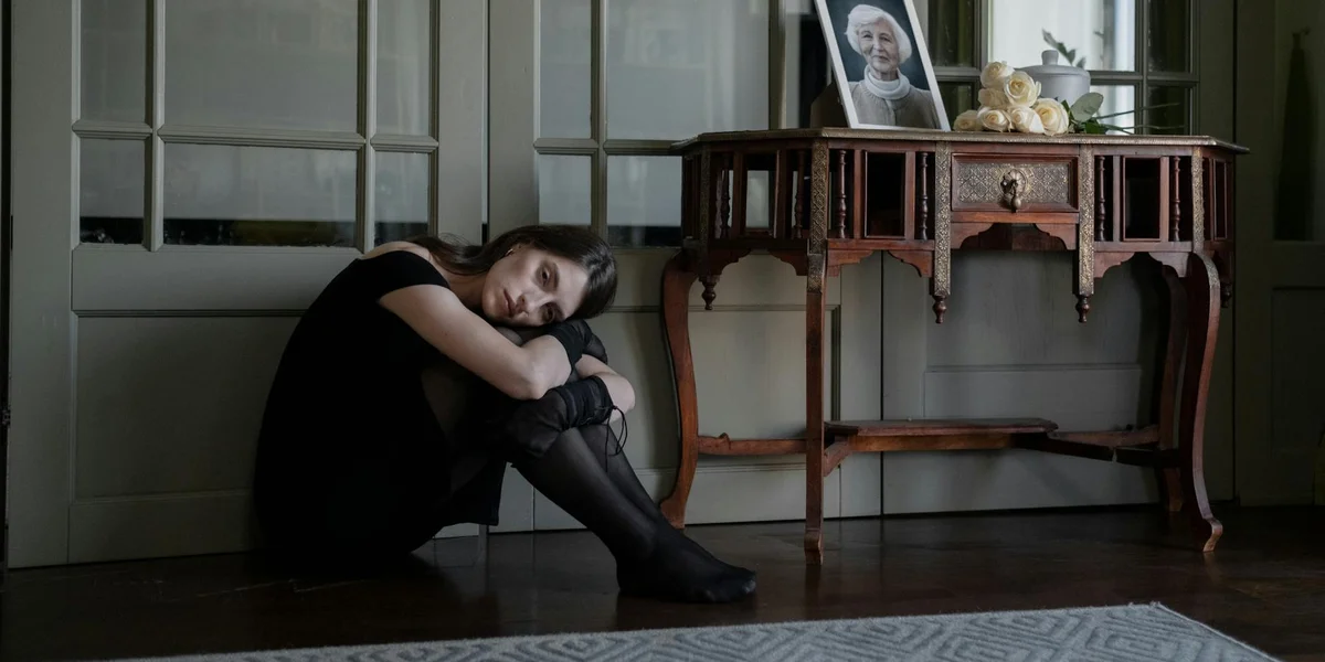 A person sits on the floor with knees drawn to their chest, resting against a wall beside a wooden console table. A framed photograph of a deceased loved one sits on the table, conveying memory and loss.