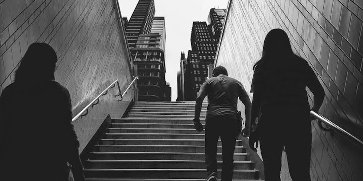 Black-and-white photograph of three silhouetted figures walking down an outdoor stairwell between tall urban buildings, suggesting tension and a sense of foreboding.