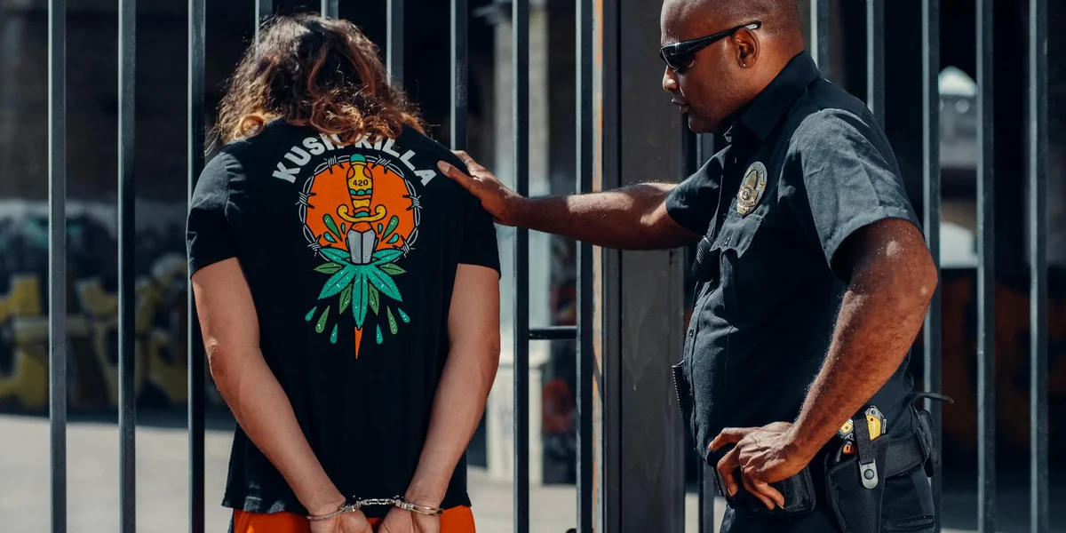 A police officer stands beside a person with hands behind their back near metal bars, suggesting a tense confrontation.