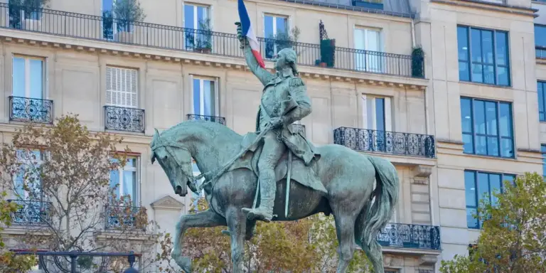 Bronze equestrian statue of a historical leader on horseback in an urban square, with a light-colored building and balconies in the background and trees in the foreground.