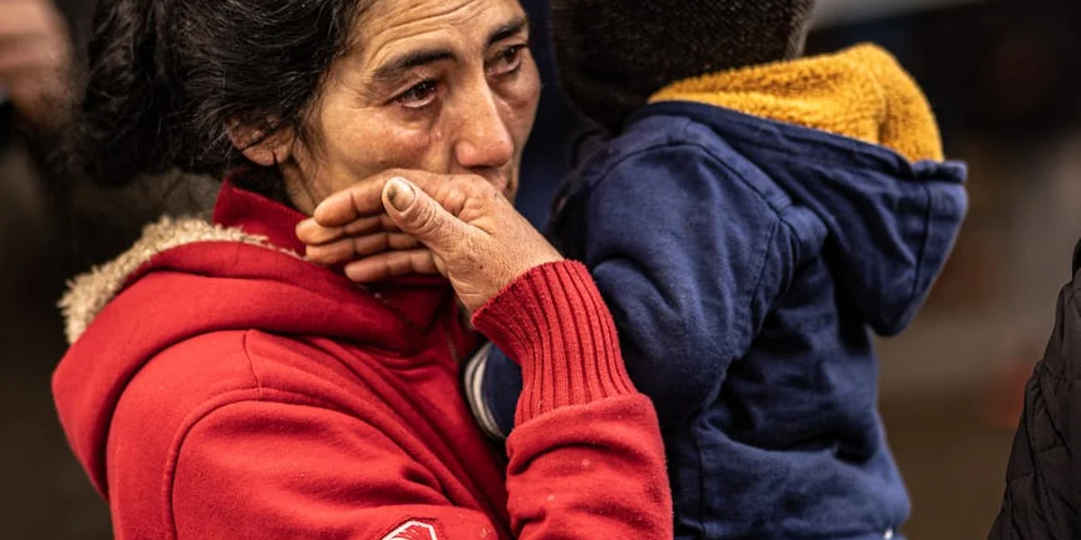 A person in a red hoodie comforts a child in a blue jacket; both look somber in a close-up moment.