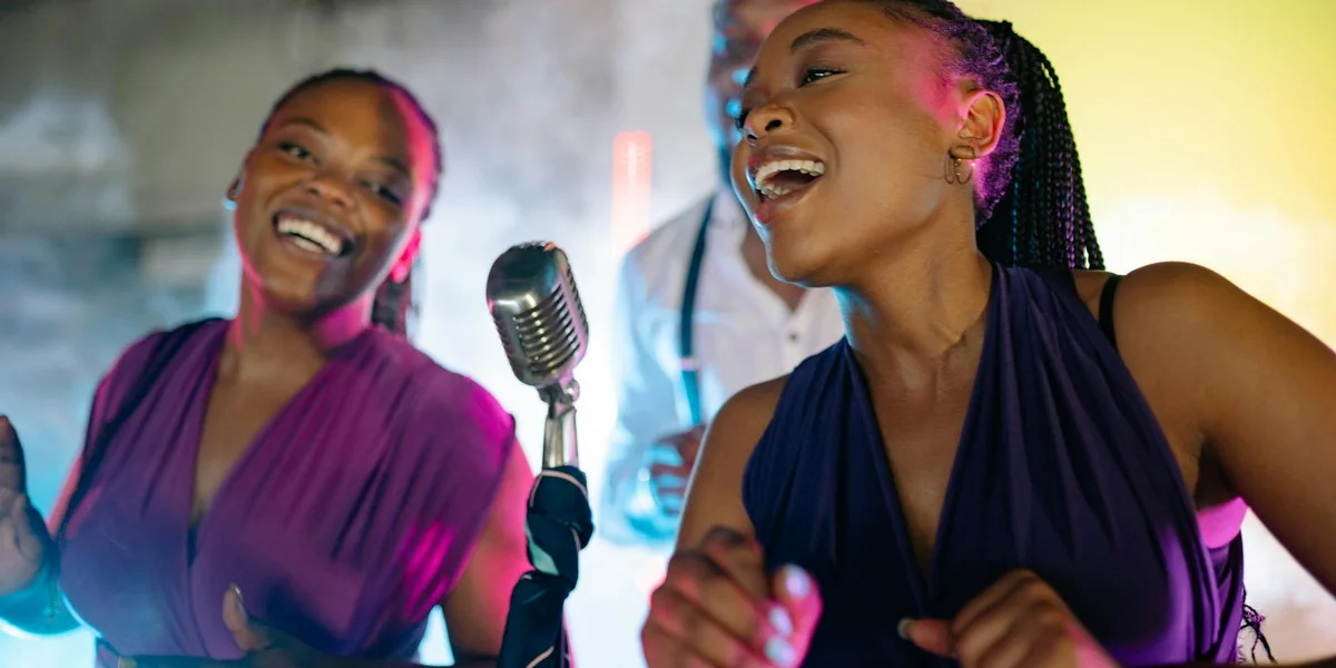 Two women singing with a vintage microphone on stage, smiling and enjoying a performance.