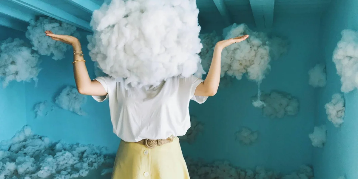 A person with a fluffy cloud for a head stands in a blue room with more cloud-like formations, arms raised