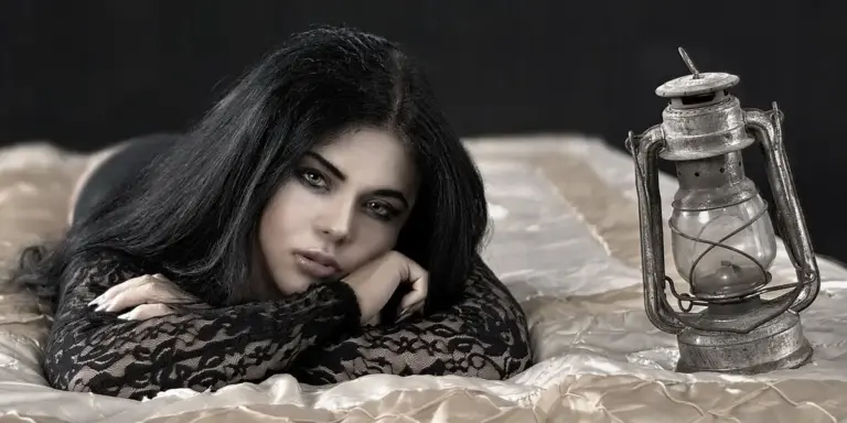 Woman with dark hair lying on a bed in a black lace outfit, next to a vintage lantern
