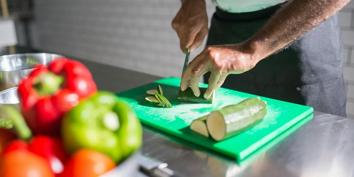 Close-up of a person slicing a cucumber on a green cutting board with colorful peppers in the foreground