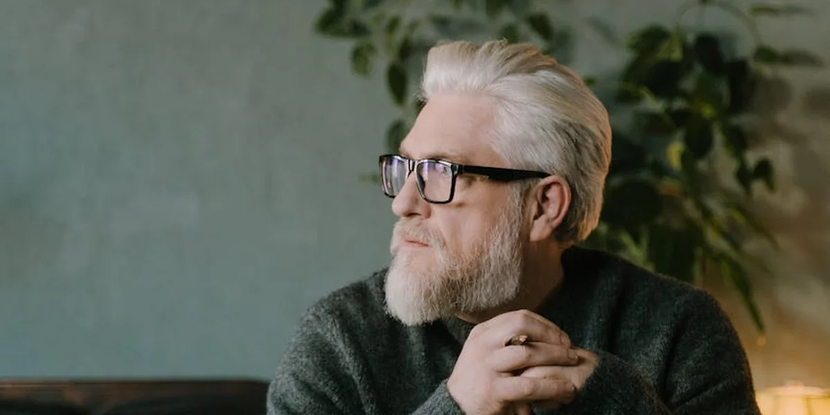 Portrait of an older man with white hair and beard wearing glasses, seated indoors, deep in thought.