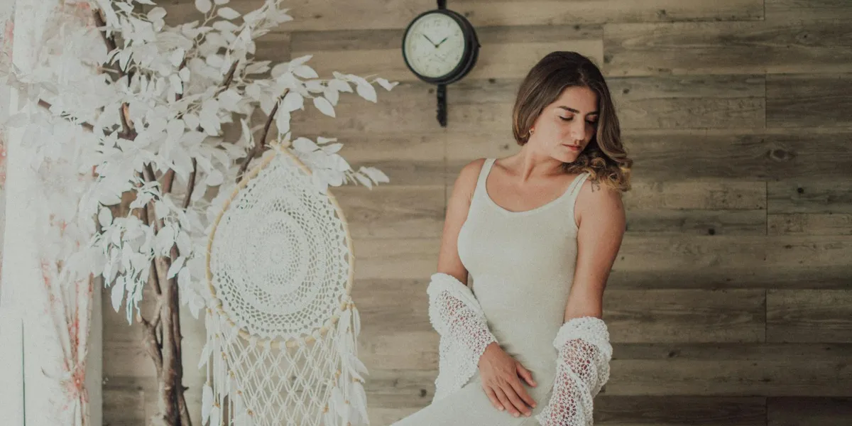 A woman in a white tank top stands indoors, looking down thoughtfully. She is beside a macrame wall hanging and a round clock on a wooden wall.