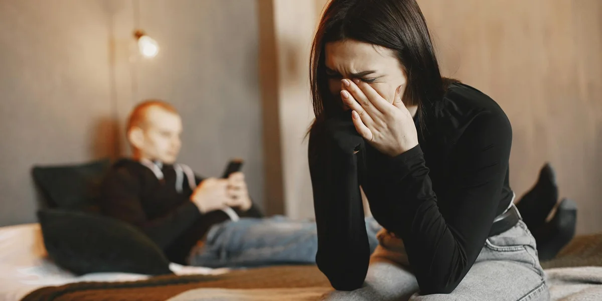 A woman in the foreground covers her face with her hands, crying, as a man sits on a couch in the background looking at a cellphone in a dim living room.