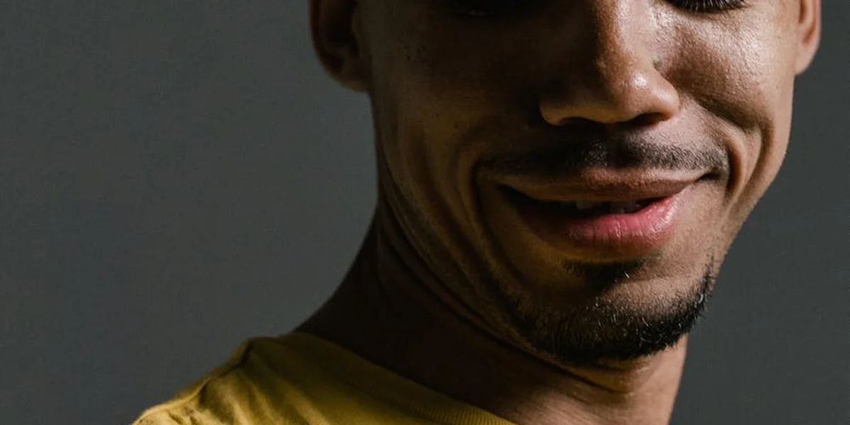Close-up of a smiling man's lower face and neck against a dark background, wearing a yellow shirt.