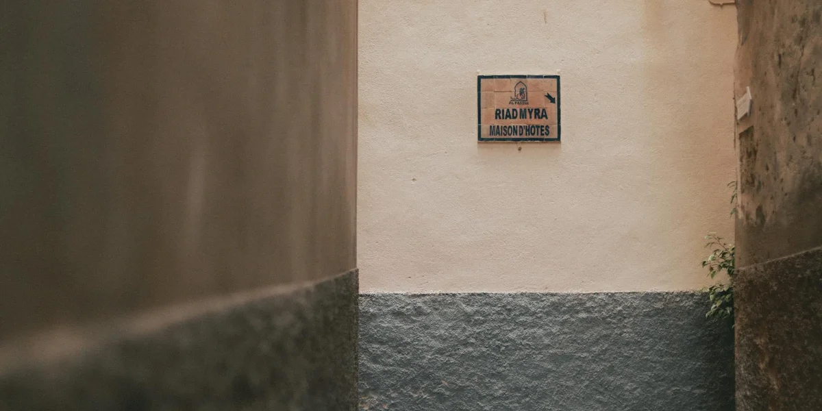 Narrow sunlit alley with tall beige walls and a blue-gray base, featuring a small sign on the wall.