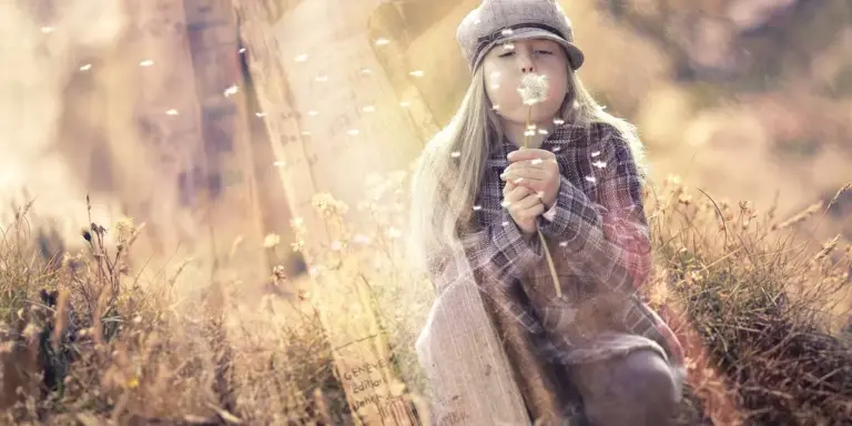 A young girl with long hair and a hat sits in a sunlit autumn field, blowing on a dandelion, with soft, dreamy light surrounding her.