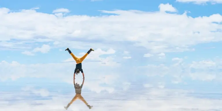 Person performing a handstand on a reflective surface under a blue sky with clouds, creating a symmetrical reflection.