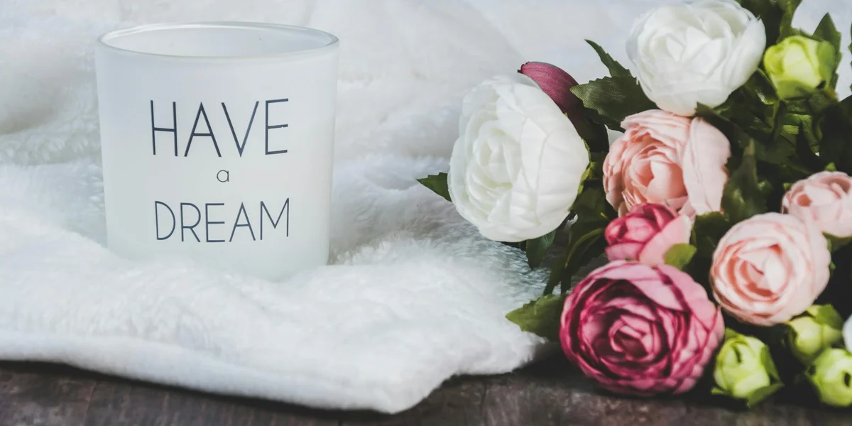 A white mug reading 'HAVE a DREAM' sits on soft white fabric beside a bouquet of pink and white roses.
