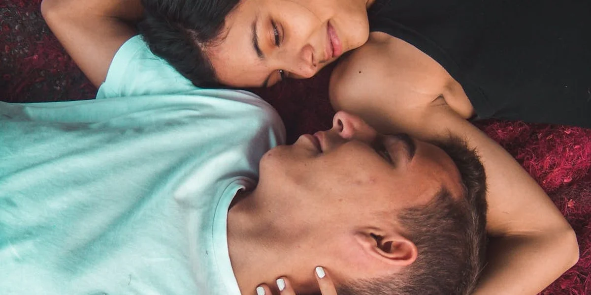 Two people lie in bed, facing each other with relaxed expressions, sharing an intimate, peaceful moment.