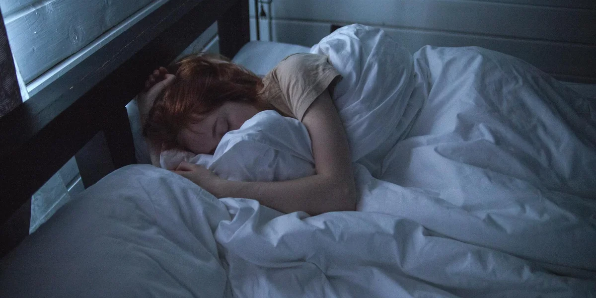 A person lies curled in bed under white sheets with their head on a pillow; the scene conveys tension and the vulnerability associated with nightmares.