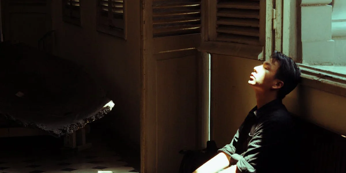 A person in a dimly lit room sits by an open doorway, looking toward a sliver of light coming from outside.