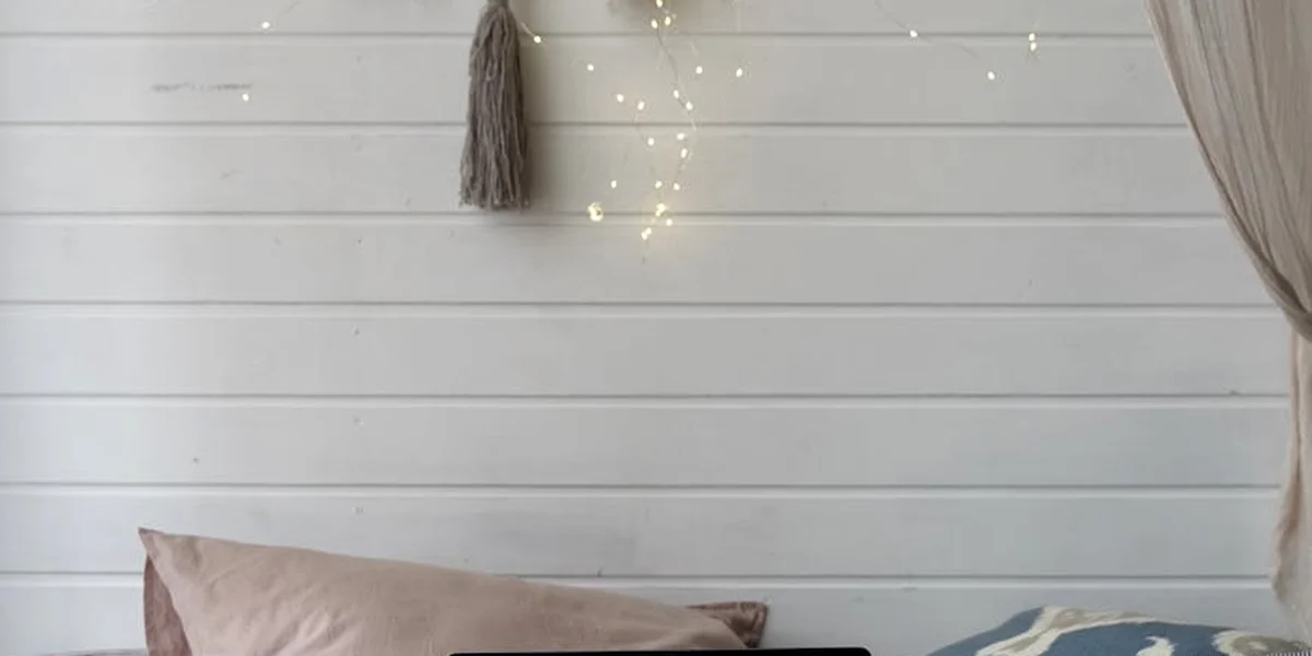 A cozy, light-colored interior featuring a pale beige cushion on a low seating surface, a decorative tassel hanging from the wall, soft string lights above, and sheer curtains at the right.
