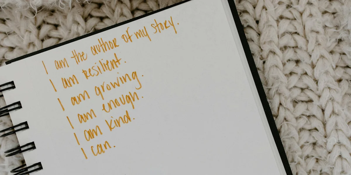 Notebook page with orange handwritten affirmations—'I am resilient. I am growing. I am kind. I can.'—on a cream knit blanket.