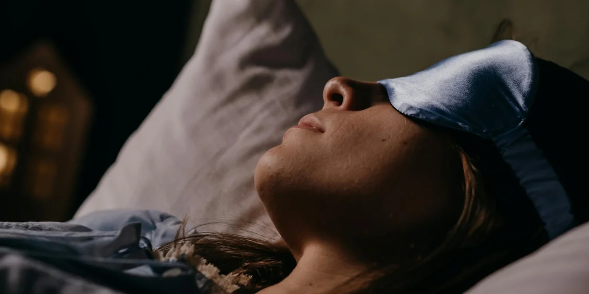 Person lying in bed wearing a sleep mask, ready to drift into sleep in a dimly lit room.