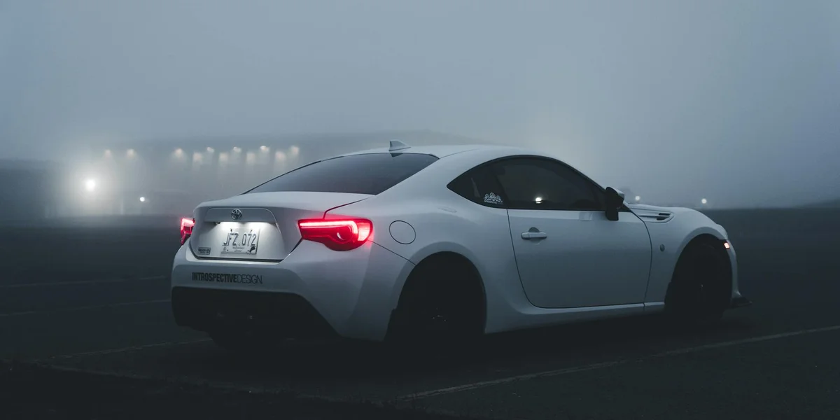 White sports car on a foggy, dark highway at night with headlights piercing through mist.