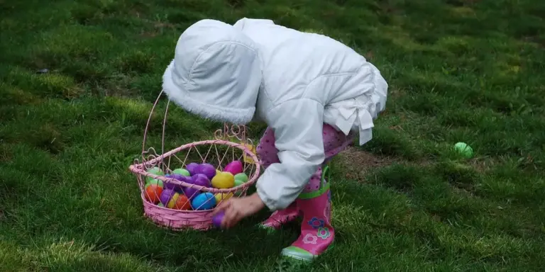 A child wearing a white hooded jacket and pink boots bends over on a grassy lawn, gathering colorful eggs into a pink wicker basket.