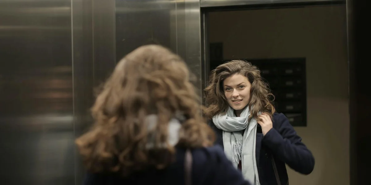 A woman in a dark coat and light scarf stands inside a metal elevator, gazing at her reflection in the doors.