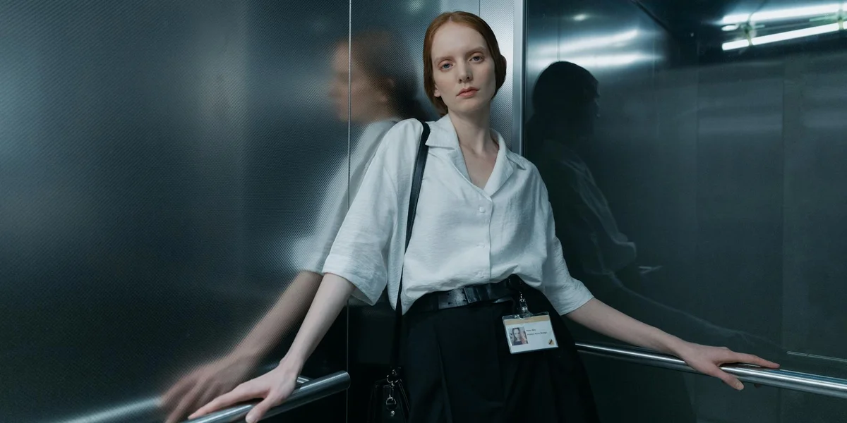 Businesswoman in an elevator, wearing a white blouse and dark trousers, leaning on the railing with a tense, wary expression.