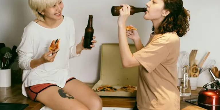 Two people in a kitchen share pizza; one drinks from a bottle while the other holds a slice, with a pizza visible on the counter.