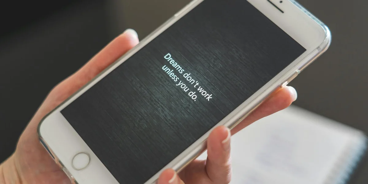 Close-up of a hand holding a smartphone with a dark screen displaying the quote 'Dreams don't work unless you do.'