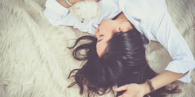 A woman with dark hair lies on a fluffy white rug, wearing a white shirt.