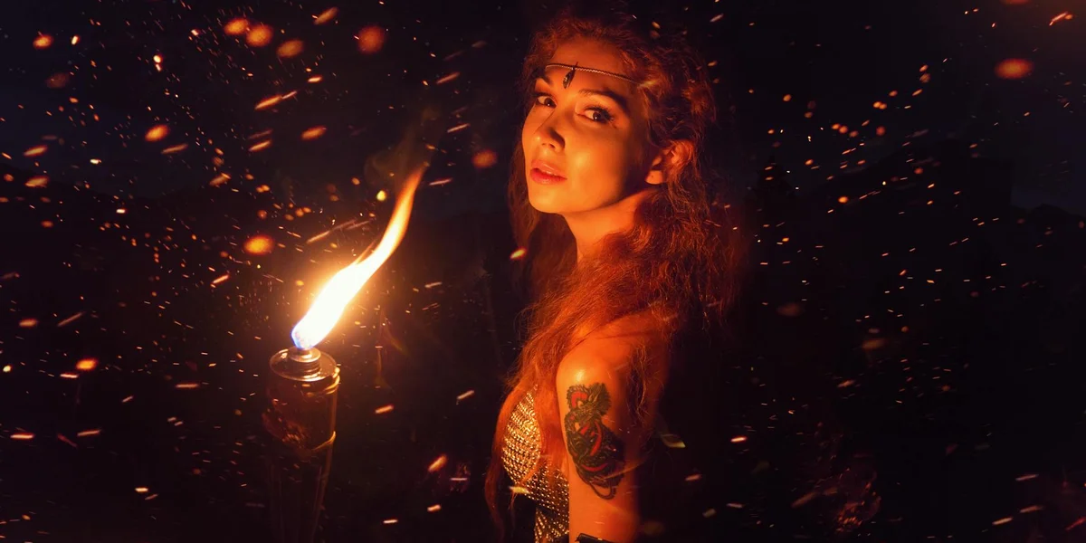 A woman stands in a dark space, holding a lit torch with sparks around, symbolizing ignition and spiritual illumination.