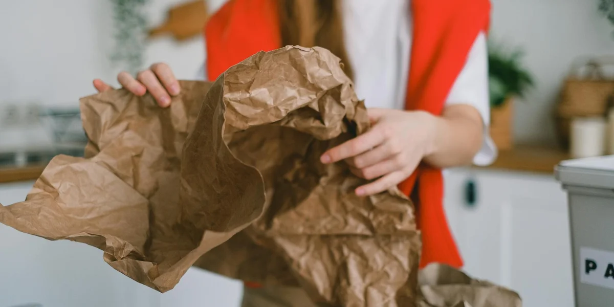 A person wearing a red top holds crumpled brown paper bags, resembling discarded trash.