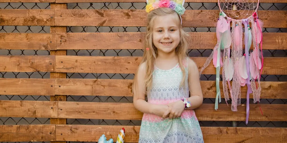 A young girl wearing a pastel dress and a colorful unicorn headband stands in front of a wooden fence, with a dreamcatcher hanging nearby.