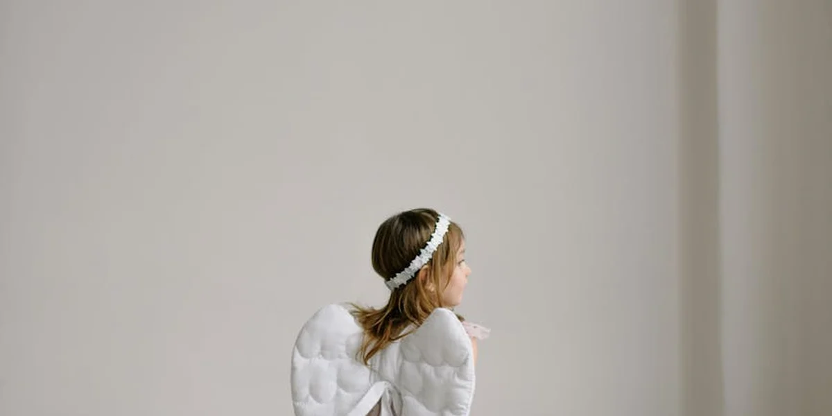 A serene woman wearing a white dress and a delicate flower headband stands against a soft, pale background, conveying a dreamlike, spiritual moment.