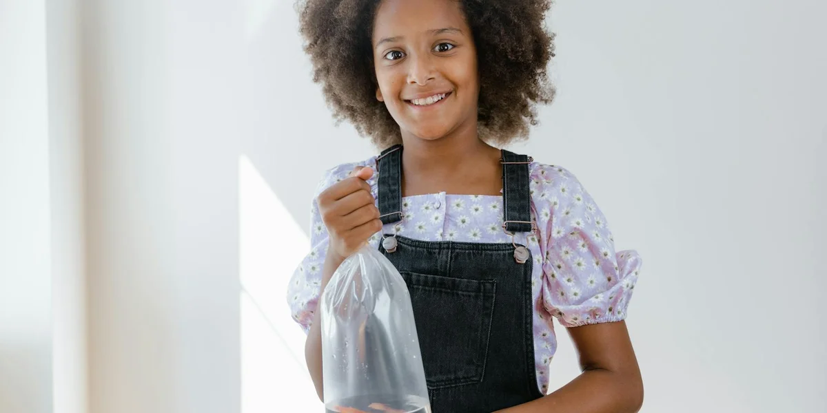 Smiling young girl with curly hair wearing a purple puff-sleeve blouse and black overalls, standing indoors and holding a plastic bag.