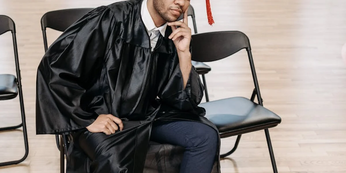A student in a graduation gown sits on a row of folding chairs, resting their chin on their hand with a worried expression.