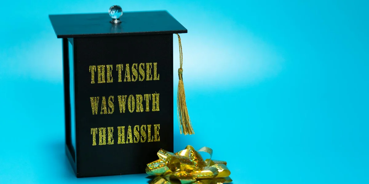 Graduation cap with a gold tassel on a blue background, featuring the inscription 'THE TASSEL WAS WORTH THE HASSLE'.
