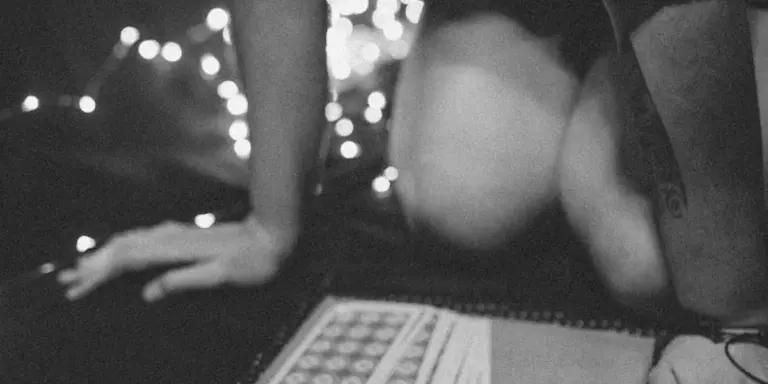 A grayscale photograph showing a person on their hands and knees beside a spiral notebook, with soft string lights in the background, suggesting contemplation and dream imagery.
