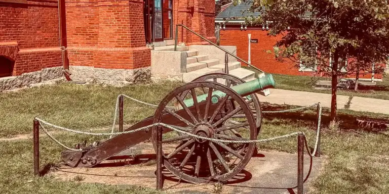 Historic cannon on a wheeled carriage displayed on a grassy area near a red-brick building, with a rope barrier around it.