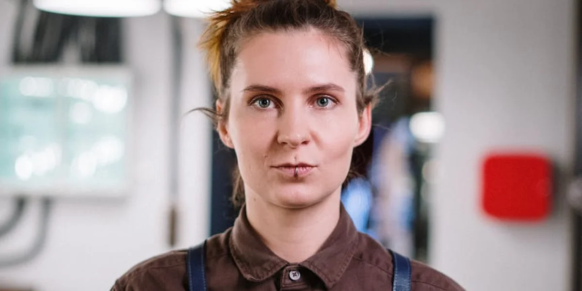 Portrait of a woman with light brown hair tied back, wearing a brown collared shirt, standing in a bright indoor setting and looking directly at the camera.