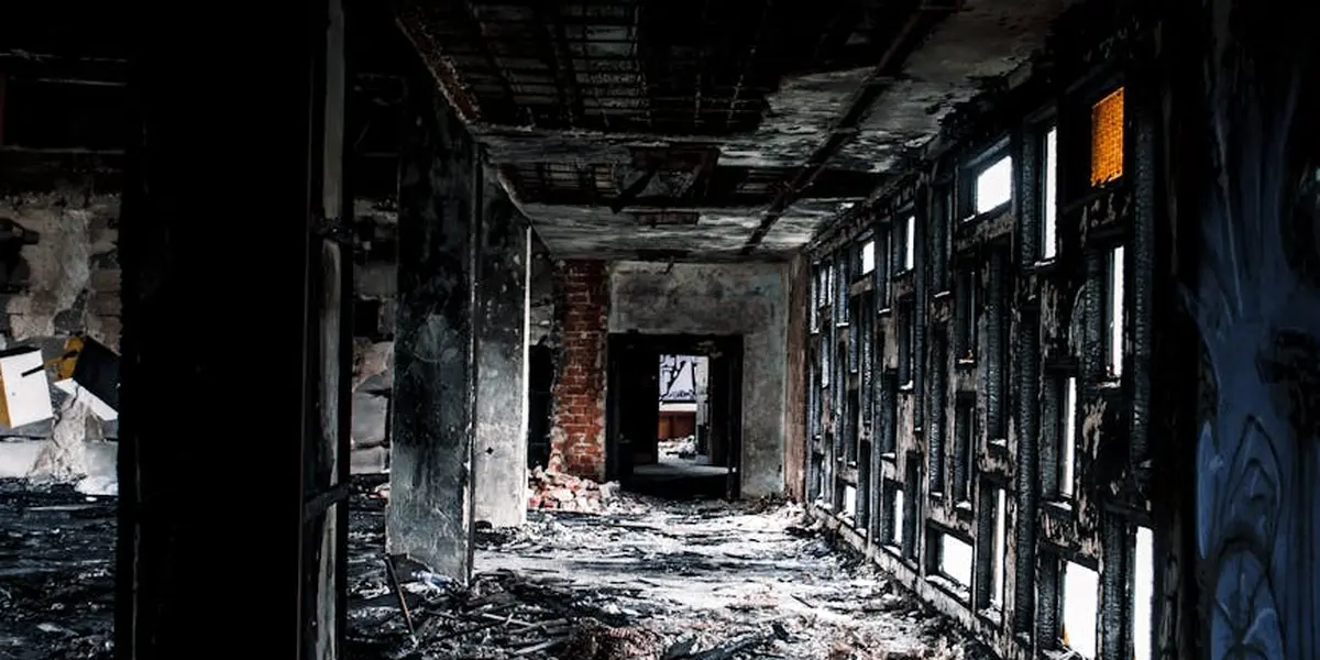 Dark, burned hallway of a house with debris on the floor and broken walls; light filters through narrow windows on the right.