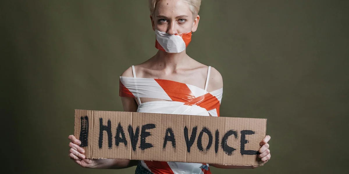 Person with taped mouth holding a cardboard sign reading 'HAVE A VOICE', symbolizing suppression of speech in dreams
