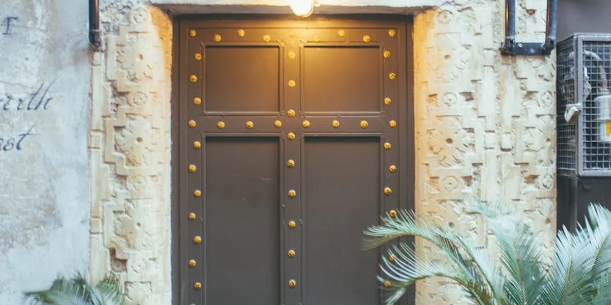 Close-up of a heavy dark wooden door with brass studs set in a stone wall, lit by a single overhead light, with tropical plants nearby.