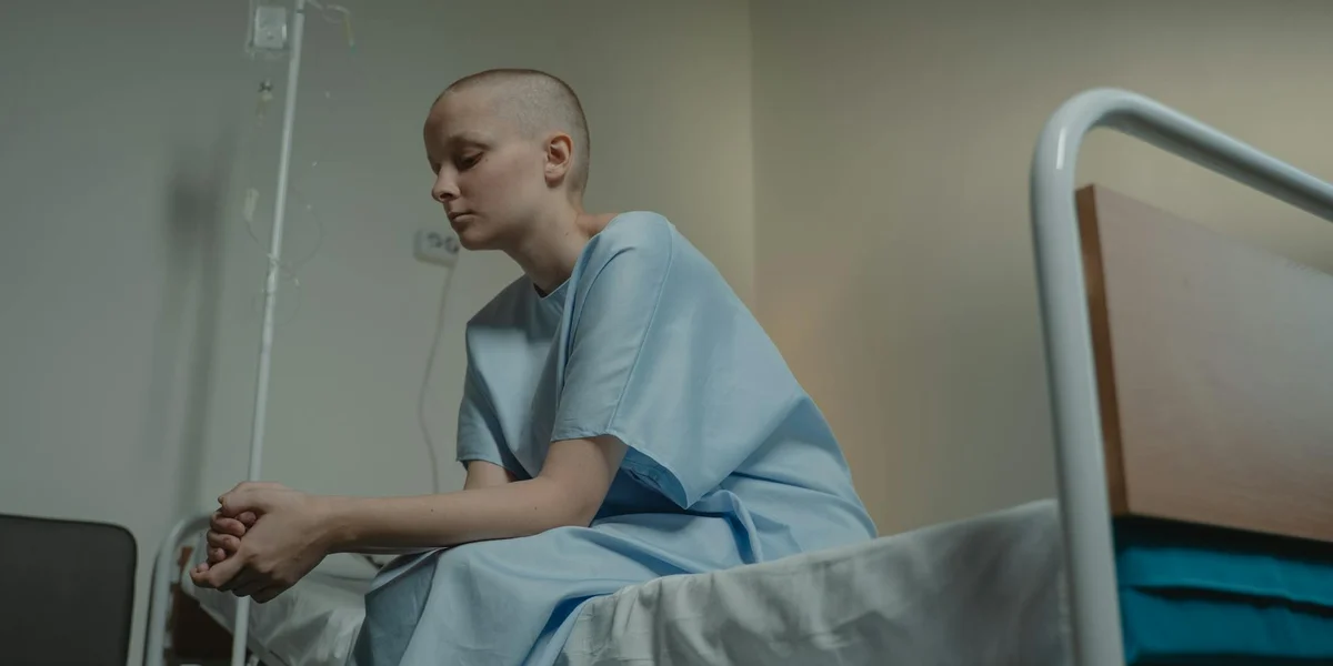 A person in a light blue hospital gown sits on the edge of a hospital bed, looking down with a contemplative expression in a quiet room.