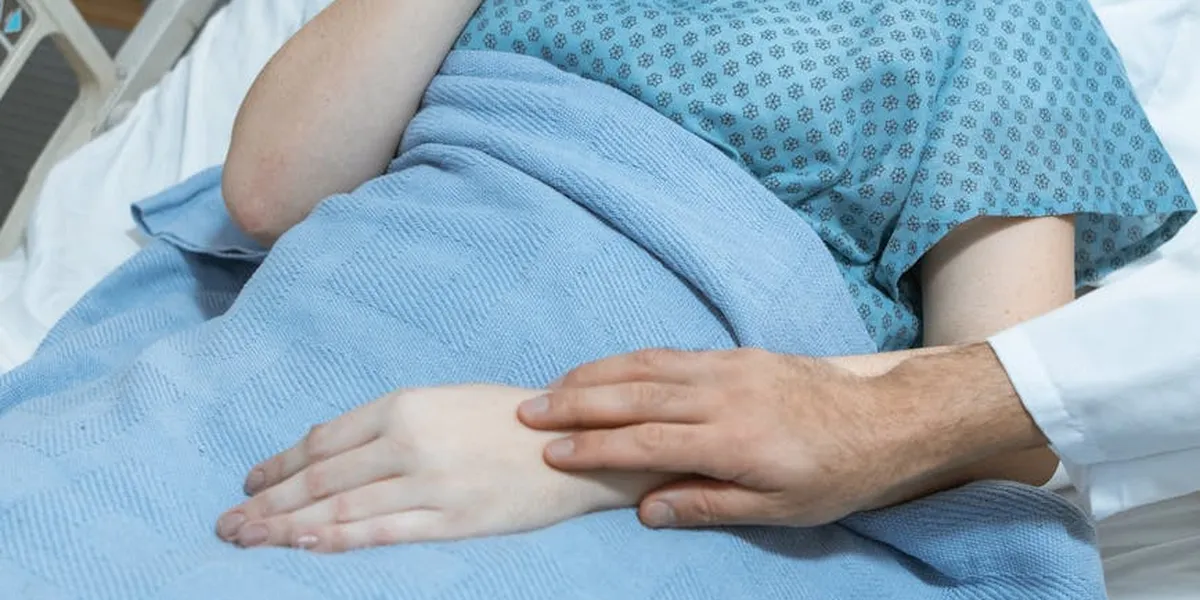 Close-up of a patient in a blue hospital gown lying in bed, with a physician's hand resting gently on the patient's hand under a blue blanket.