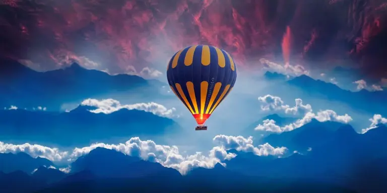 A bright orange and blue hot air balloon floating among fluffy white clouds in a clear blue sky above distant mountains.