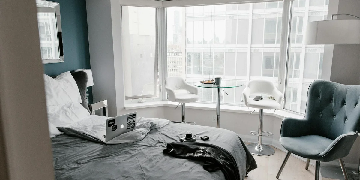 A modern hotel room with a bed, a laptop resting on the bed, a teal armchair, and a window seating area with white chairs and a small round table.
